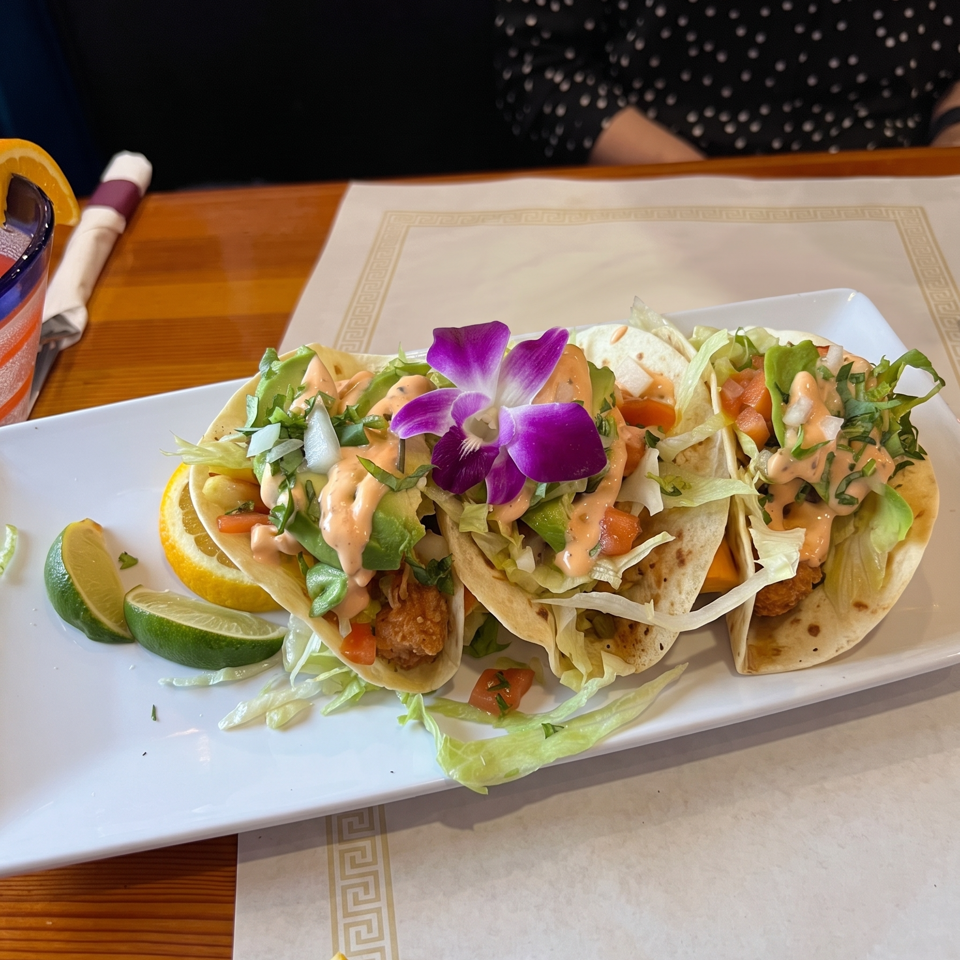 Three soft tacos topped with lettuce, avocado, tomatoes, creamy sauce, herbs, and purple orchids, served with lime and lemon wedges.