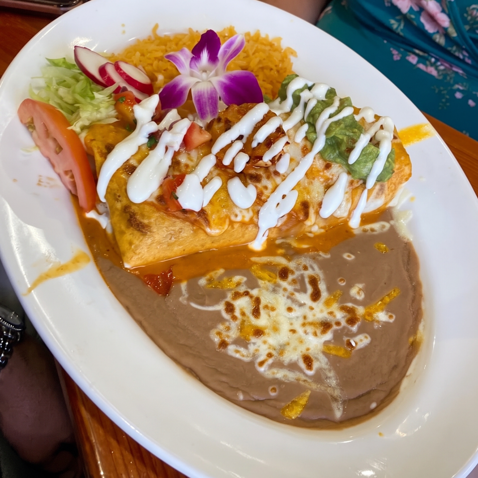 Mexican enchilada plate with yellow rice, refried beans, guacamole, vegetables, sour cream, and a purple flower garnish.