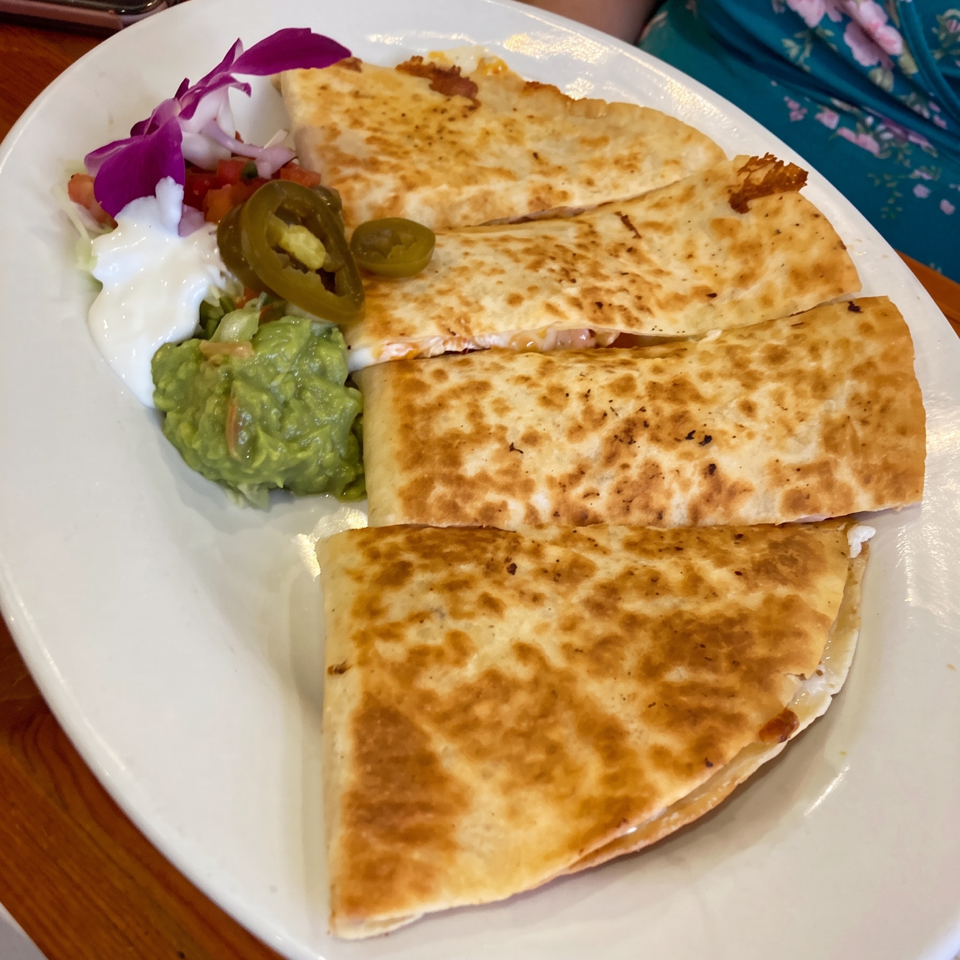 Golden-brown quesadilla wedges served with guacamole, sour cream, jalapeños, salsa, and edible purple flowers on a white plate.