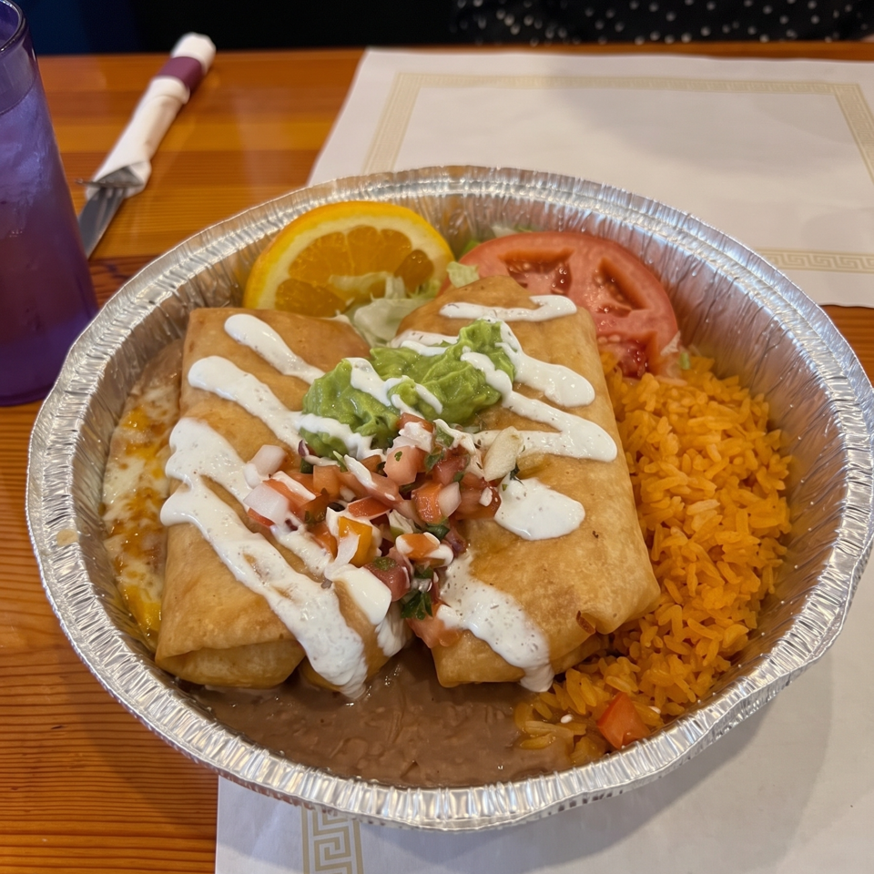 Two fried burritos topped with guacamole, pico de gallo, and cream sauce, served with Mexican rice, salad, and an orange slice.